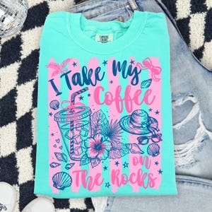 May include: A turquoise Comfort Colors t-shirt with a graphic that says "I take my coffee on the rocks." The design includes a coffee drink, flowers, a hat, sunglasses, and seashells in pink and blue.