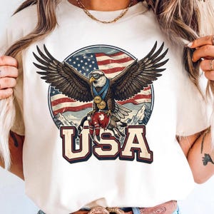 May include: Cream-colored t-shirt featuring a graphic of an eagle with outstretched wings, wearing goggles and a medal. The eagle is perched atop a hockey jersey with a maple leaf. The background includes the American flag, mountains, and the word "USA".