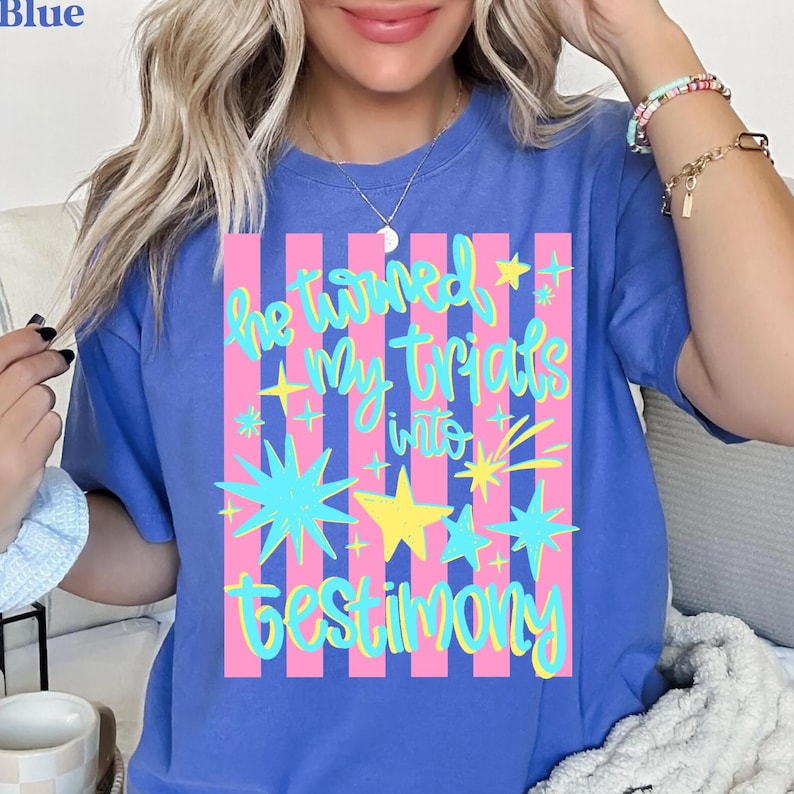 May include: Blue t-shirt with a colorful graphic. The graphic features the text "He turned My trials into Testimony" in a whimsical font, with stars and stripes in pink, yellow, and turquoise. The shirt is a solid blue color.