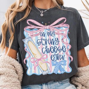 May include: Dark grey t-shirt with the text "in my string cheese era". The graphic includes a string cheese stick, pink and blue bows, and a pastel striped background.