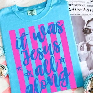 May include: A light blue t-shirt with the text "it was Jesus all along" in a cursive font, with pink vertical stripes. The shirt has a crew neck and short sleeves. The Comfort Colors brand tag is visible.