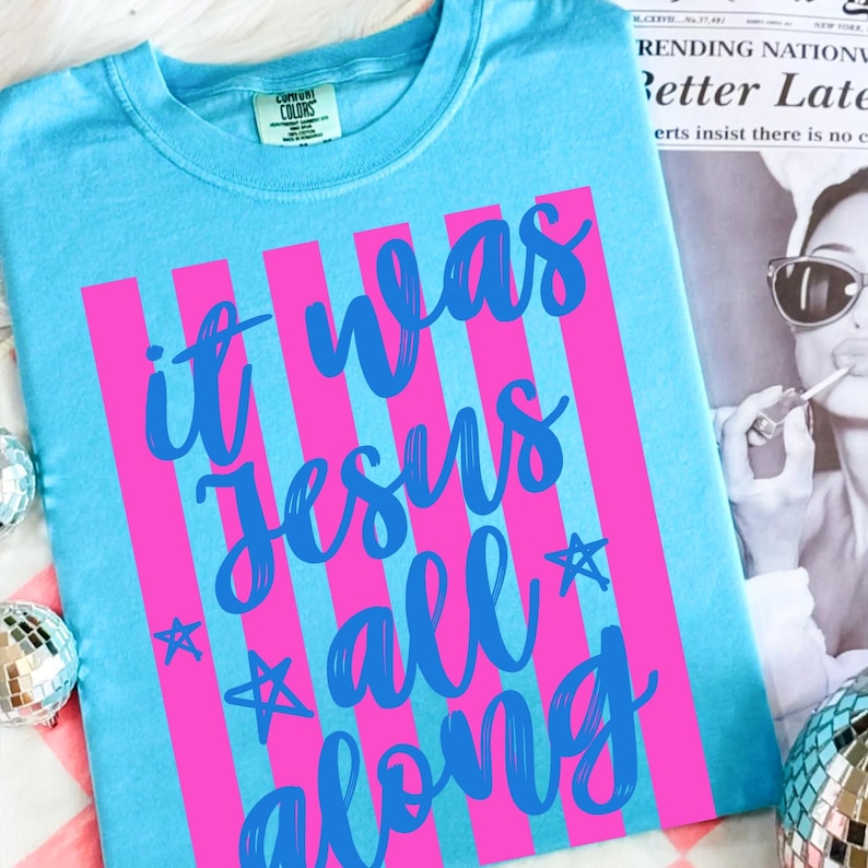 May include: A light blue t-shirt with the text "it was Jesus all along" in a cursive font, with pink vertical stripes. The shirt has a crew neck and short sleeves. The Comfort Colors brand tag is visible.