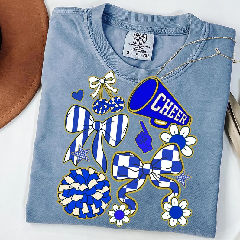 May include: A light blue Comfort Colors t-shirt featuring cheerleading-themed graphics. The design includes a blue megaphone with the word "CHEER", bows, pom-poms, and flowers in blue and white. The shirt has a crew neck and short sleeves.