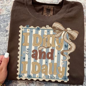 May include: A brown Comfort Colors t-shirt featuring a decorative design with the words "I Dilly and I Dally" in a vintage font. The design includes a bow and floral accents on a blue and white striped background.