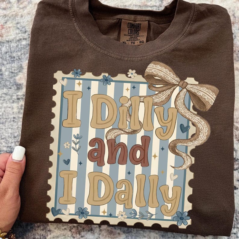 May include: A brown Comfort Colors t-shirt featuring a decorative design with the words "I Dilly and I Dally" in a vintage font. The design includes a bow and floral accents on a blue and white striped background.