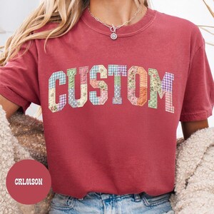 May include: Crimson-colored t-shirt with the word "CUSTOM" in a patchwork design. The letters are made of various floral and patterned fabrics. The shirt has short sleeves and a crew neck.