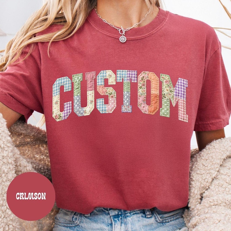 May include: Crimson-colored t-shirt with the word "CUSTOM" in a patchwork design. The letters are made of various floral and patterned fabrics. The shirt has short sleeves and a crew neck.