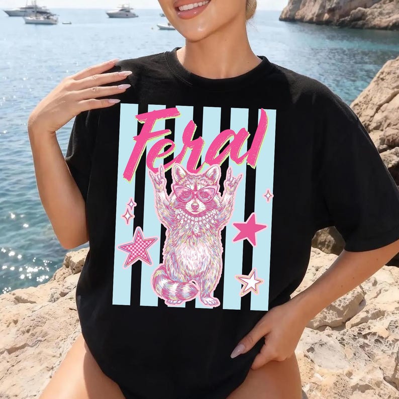 Pode incluir: Camiseta preta com um desenho rosa e branco de um guaxinim usando &oacute;culos de sol e um colar, com a palavra "Feral" acima. O guaxinim est&aacute; fazendo um gesto de rock and roll com as m&atilde;os, em um fundo de listras azuis e estrelas cor-de-rosa.