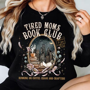 Può includere: Felpa nera con la scritta "TIRED MOMS BOOK CLUB" sopra un'immagine ovale di uno skyline, libri e una tazza di caffè. Il testo "RUNNING ON COFFEE, CHAOS AND CHAPTERS" è in basso. La felpa ha un girocollo.