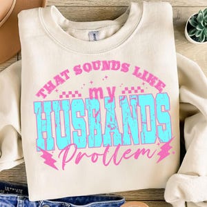 May include: Cream-colored sweatshirt with the text "THAT SOUNDS LIKE my HUSBANDS Problem" in pink and turquoise. The design includes lightning bolt graphics and a retro font. The sweatshirt is folded, with a glimpse of denim jeans visible.