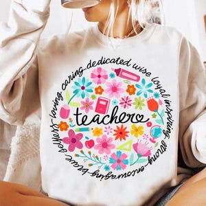 May include: White sweatshirt with a colorful circle design featuring the word "teacher" surrounded by flowers, school supplies, and the words "smart - loving - caring - dedicated - wise - loyal - inspiring - brave - strong - kind -  true".