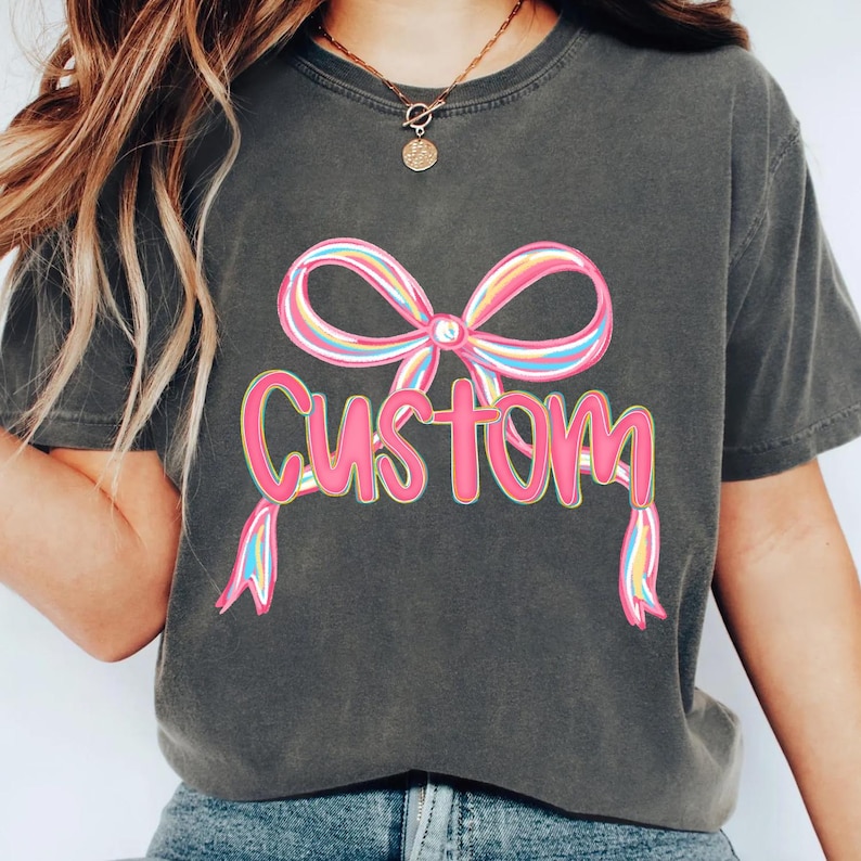 May include: Dark grey t-shirt featuring a colourful bow graphic and the word "Custom" in pink. The bow has stripes of pink, yellow, blue, and green. The shirt is a casual style.