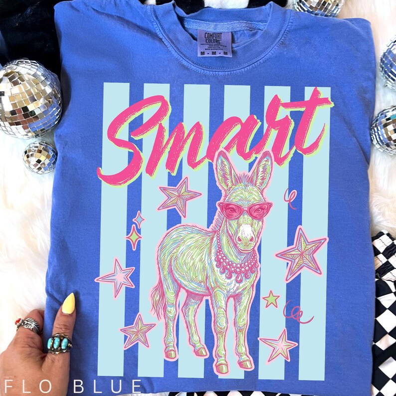 Pode incluir: Camiseta azul Flo com um gr&aacute;fico de um burro usando &oacute;culos de sol e um colar de p&eacute;rolas, com a palavra "Smart" em escrita rosa. O design inclui listras azuis claras e estrelas.