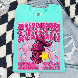 May include: A turquoise t-shirt with a pink and white graphic. The graphic features the word "KNIGHTS" in pink with white stars, a pink helmet, and the words "SCHOOL NAME". The shirt is made of a soft material.