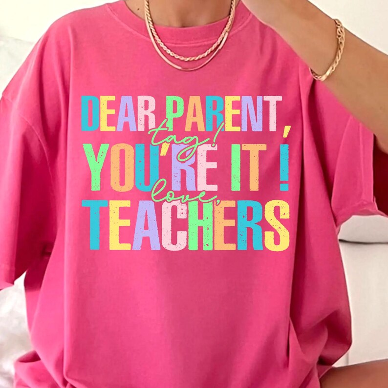 May include: A bright pink t-shirt with the text "DEAR PARENT, YOU'RE IT! LOVE TEACHERS" in a colorful, blocky font. The shirt has a crew neck and short sleeves. The text is in shades of blue, green, yellow, and purple.