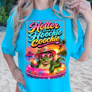May include: A turquoise t-shirt features a colorful graphic of an alligator in sunglasses and a hat, floating in a pool with a cocktail. The text reads "Hotter Than a Hoochie Coochie" in a retro font, with a tropical sunset background.