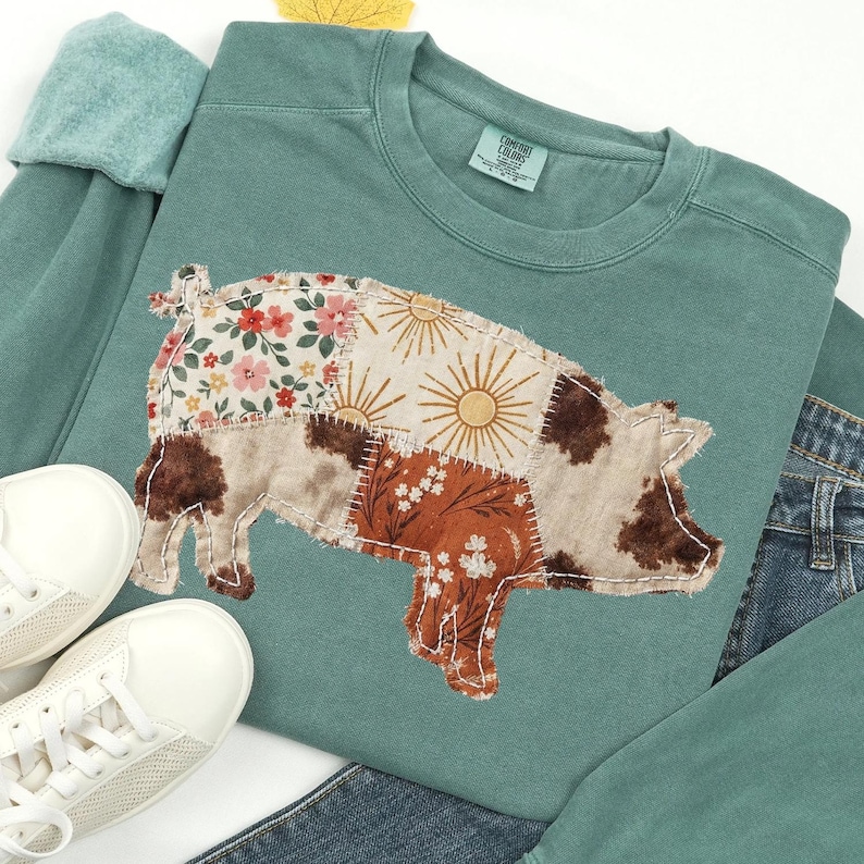 May include: A teal sweatshirt featuring a patchwork pig design. The pig is made of various patterned fabrics, including floral, sunburst, and cowhide prints. White sneakers and denim jeans are also visible.
