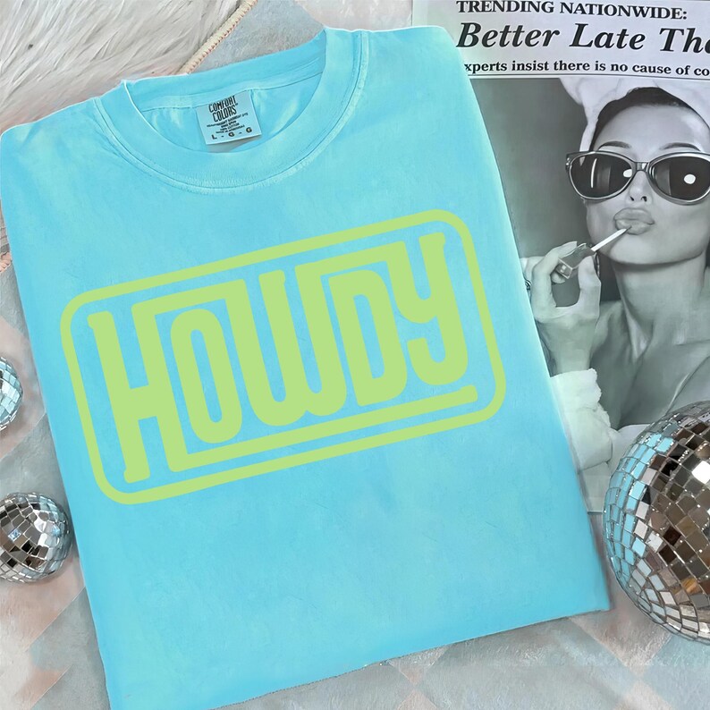May include: A light blue Comfort Colors t-shirt with the word "HOWDY" in a neon green, outlined font. The shirt is folded and displayed with a newspaper clipping and a disco ball in the background.