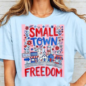 May include: Light blue t-shirt with a graphic design featuring the words "SMALL TOWN FREEDOM" in red and blue. The design includes illustrations of buildings, flowers, and a road, with a pink striped background.