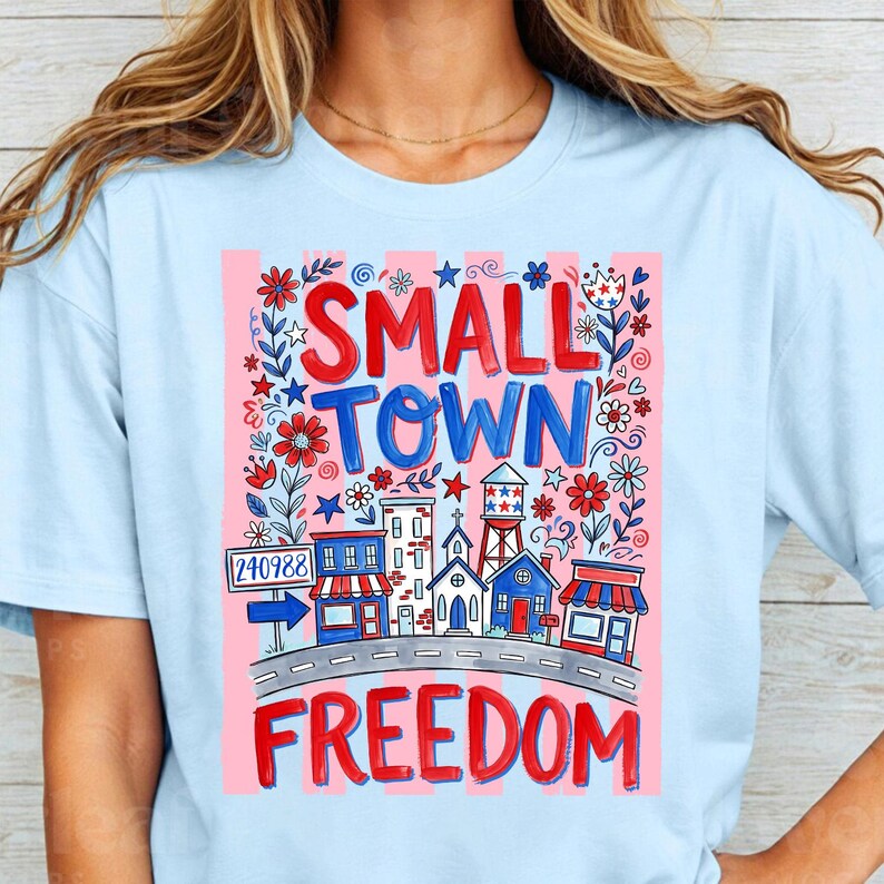 May include: Light blue t-shirt with a graphic design featuring the words "SMALL TOWN FREEDOM" in red and blue. The design includes illustrations of buildings, flowers, and a road, with a pink striped background.