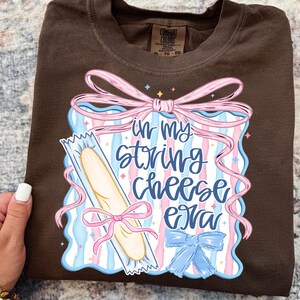 May include: A brown t-shirt with the phrase "in my string cheese era" in a graphic design. The design includes a string cheese stick, pink and blue ribbons, and a blue bow. The shirt has a crew neck and short sleeves.