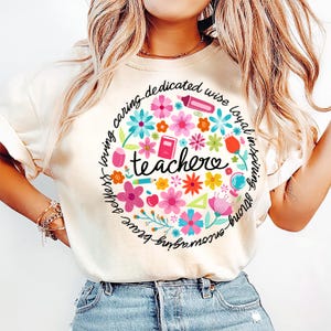 May include: A white t-shirt with a circle design featuring colorful flowers, school supplies, and the word "teacher" in the center. The circle is surrounded by the words "strong, brave, kind, loving, caring, dedicated, wise, loyal, inspiring".