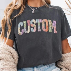 May include: Dark gray t-shirt with the word "CUSTOM" in colorful patchwork letters. The letters are made of various floral and patterned fabrics, creating a unique and eye-catching design. The shirt has a casual, vintage look.