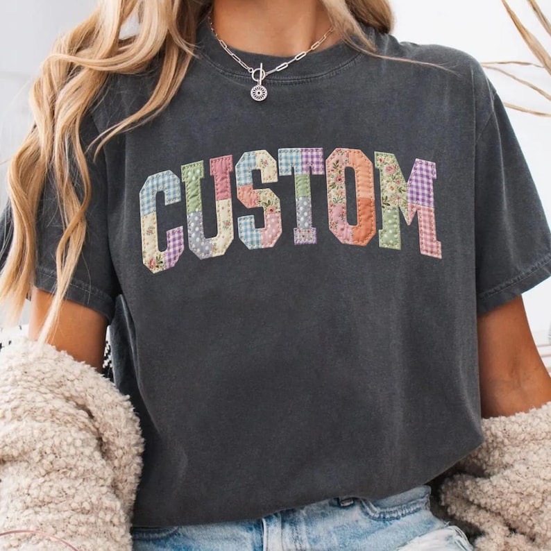May include: Dark gray t-shirt with the word "CUSTOM" in colorful patchwork letters. The letters are made of various floral and patterned fabrics, creating a unique and eye-catching design. The shirt has a casual, vintage look.