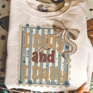 May include: Cream-colored sweatshirt with the phrase "I Dilly and I Dally" in a vintage-style design. The graphic includes a decorative frame, floral accents, and a bow.