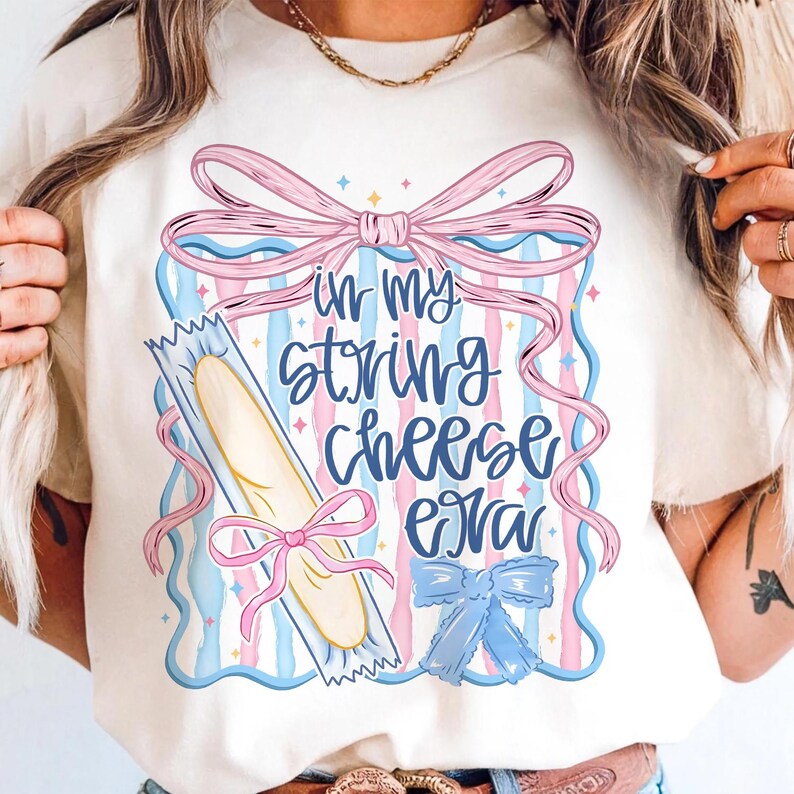 May include: Cream t-shirt featuring the text "In My String Cheese Era" in blue script. The design includes a string cheese stick, pink and blue ribbons, and a bow. The shirt has a casual, playful design.