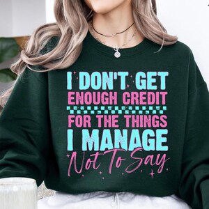 May include: Dark green sweatshirt with the text "I DON'T GET ENOUGH CREDIT FOR THE THINGS I MANAGE Not To Say" in pink and light blue. The sweatshirt has a crew neck and long sleeves.