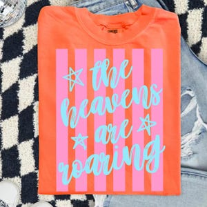 May include: Bright orange t-shirt with pink vertical stripes and the text "the heavens are roaring" in light blue script. The shirt has a crew neck and is made of a soft material. The shirt is displayed on a denim background.