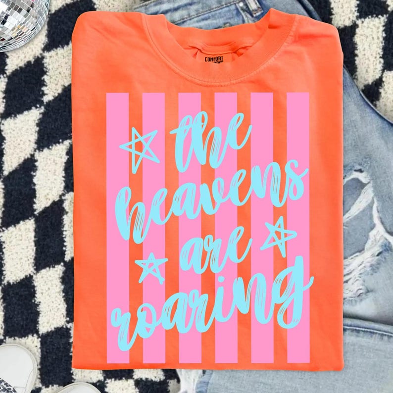 May include: Bright orange t-shirt with pink vertical stripes and the text "the heavens are roaring" in light blue script. The shirt has a crew neck and is made of a soft material. The shirt is displayed on a denim background.