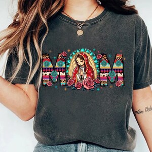 May include: Dark gray t-shirt with the word "MOM" in a colorful, patterned design. The letter "O" features an illustration of a praying figure with a halo, surrounded by roses. The shirt is a casual style.