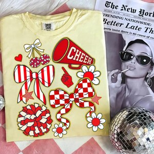 May include: A light yellow t-shirt with red and white cheerleading-themed graphics, including bows, flowers, a megaphone with the word "CHEER", and a pom-pom. A newspaper and a disco ball are in the background.