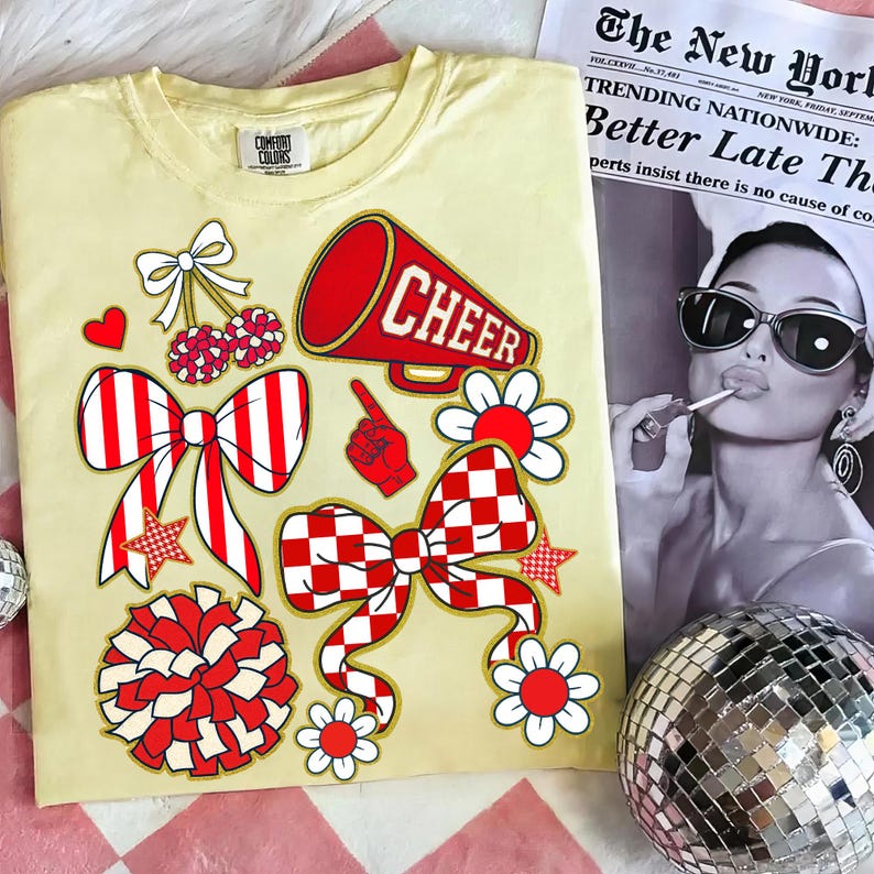 May include: A light yellow t-shirt with red and white cheerleading-themed graphics, including bows, flowers, a megaphone with the word "CHEER", and a pom-pom. A newspaper and a disco ball are in the background.