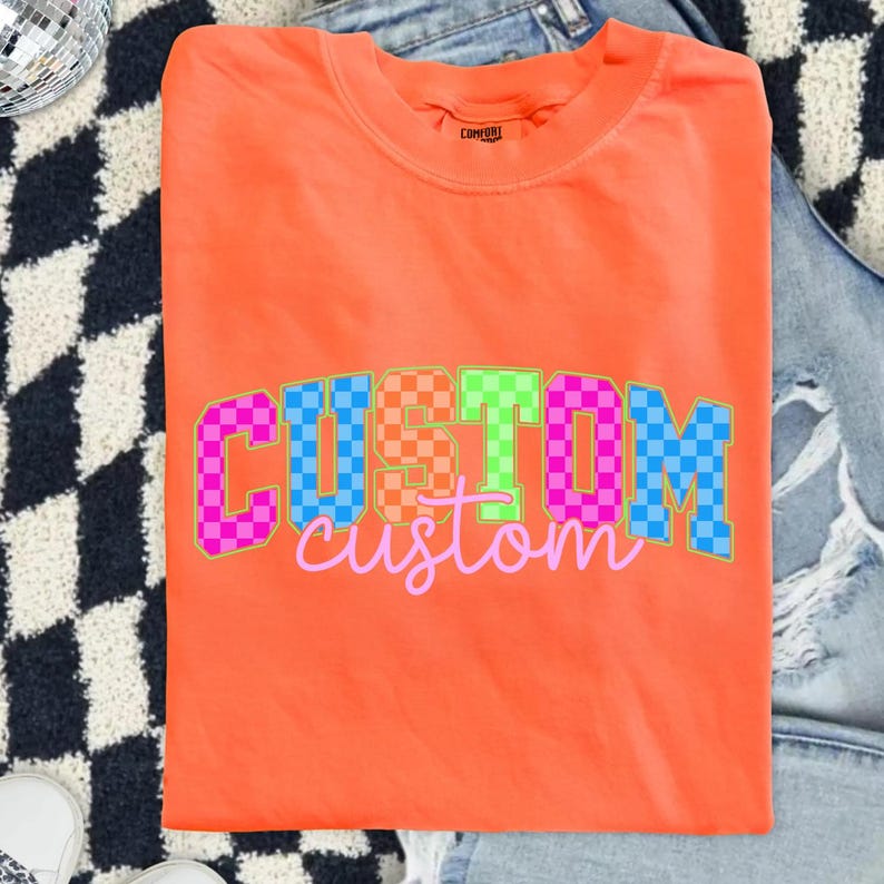 May include: Bright orange crew neck t-shirt with the word "CUSTOM" in a checkered pattern across the front. The word "custom" is written in a cursive font below the word "CUSTOM". The shirt is folded on top of a pair of blue jeans.