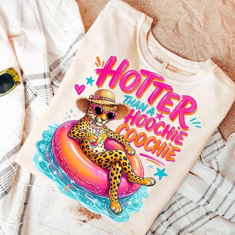 K&ouml;nnte beinhalten: Cremefarbenes Sweatshirt mit einer Grafik eines Geparden mit Sonnenbrille und Strohhut, der auf einem rosa Schwimmring schwimmt. Der Text "Hotter than a Hoochie Coochie" ist in Pink und Orange geschrieben. Das Sweatshirt liegt auf einem wei&szlig;en Handtuch und Sand.