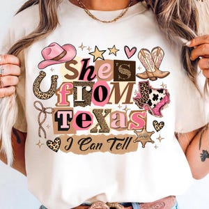 May include: Cream-colored t-shirt with a graphic design featuring the text "She's From Texas I Can Tell." The design includes a pink cowboy hat, boots, a horseshoe, and the state of Texas, all with a Western theme.