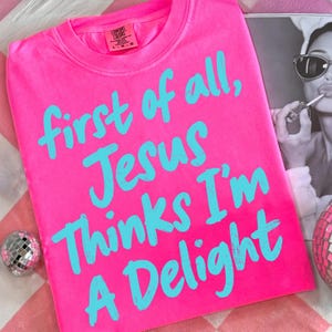 May include: A bright pink t-shirt with the text "first of all, Jesus thinks I'm a delight" in turquoise. The shirt is made of a soft material and has a crew neck. A black and white photo is in the background.