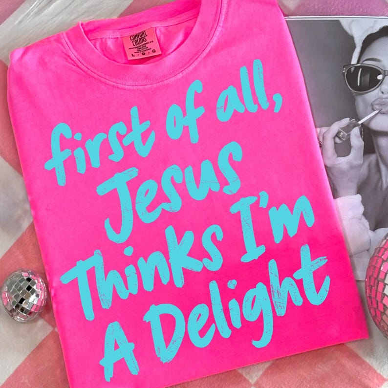 May include: A bright pink t-shirt with the text "first of all, Jesus thinks I'm a delight" in turquoise. The shirt is made of a soft material and has a crew neck. A black and white photo is in the background.
