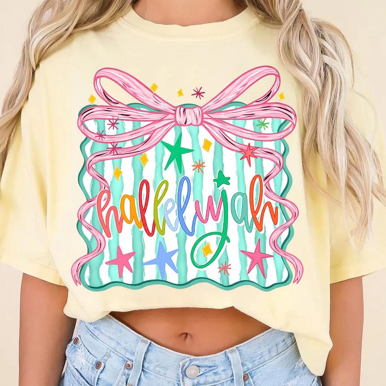 May include: A light yellow t-shirt with a colorful graphic. The graphic features a pink bow, a teal and white striped rectangle, and the word "hallelujah" in rainbow colors. Stars and confetti-like shapes surround the design.