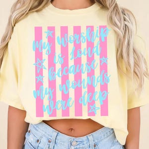 May include: A light yellow t-shirt with the text "My Worship is loud because my wounds were deep" in light blue script. The text is set against pink vertical stripes and star accents.