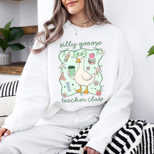 May include: White sweatshirt with a graphic of a cartoon goose holding a coffee cup, surrounded by school-themed illustrations. The text "silly goooose teacher club" is printed above and below the goose design. The design is framed by a green gingham pattern.