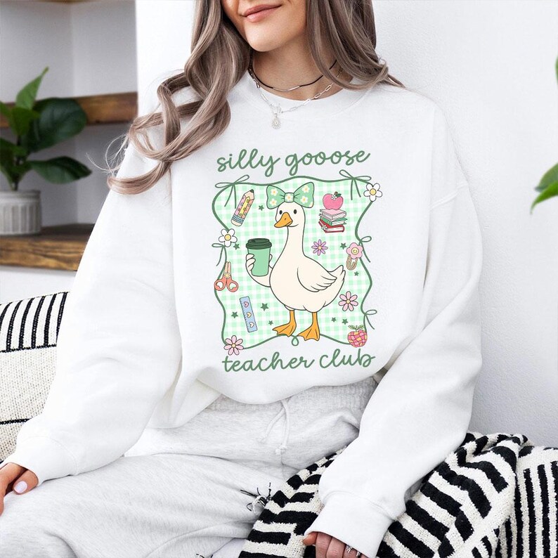 May include: White sweatshirt with a graphic of a cartoon goose holding a coffee cup, surrounded by school-themed illustrations. The text "silly goooose teacher club" is printed above and below the goose design. The design is framed by a green gingham pattern.