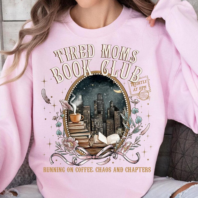 May include: Light pink sweatshirt with the text "TIRED MOMS BOOK CLUB" above an oval graphic of a cityscape, coffee cup, and books. The graphic also includes the text "RUNNING ON COFFEE. CHAOS AND CHAPTERS".