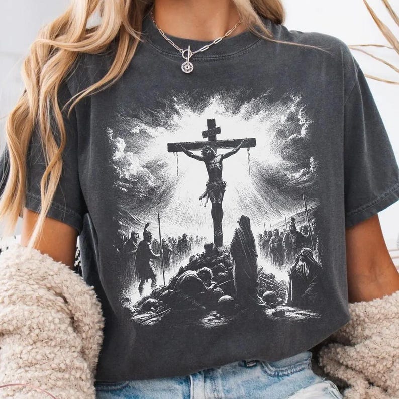 May include: Dark gray t-shirt featuring a detailed black and white illustration of the crucifixion of Jesus Christ. The graphic depicts Jesus on the cross, surrounded by figures and a dramatic sky.