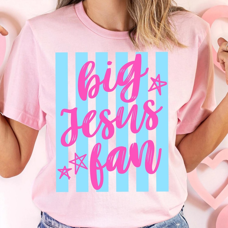 May include: A light pink t-shirt with the words "big Jesus fan" in bright pink script. The text is set against a backdrop of vertical light blue stripes, with star accents. The shirt is a casual wear item.