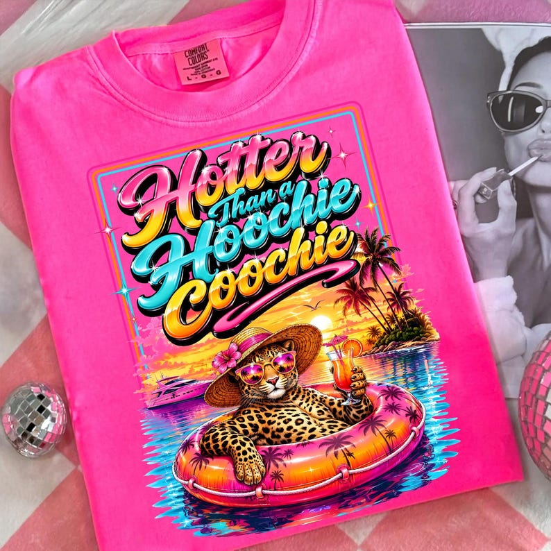 May include: Bright pink t-shirt with a graphic that reads "Hotter Than a Hoochie Coochie." The graphic features a leopard wearing sunglasses and a hat, floating in a pool with a cocktail. Palm trees and a sunset are in the background.