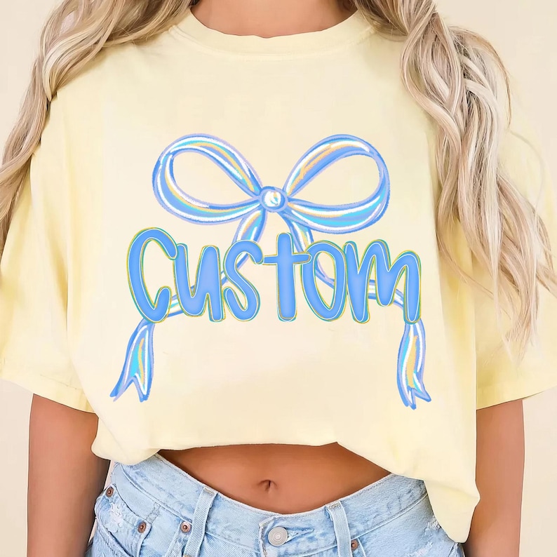 May include: A pale yellow t-shirt featuring a blue bow design and the word "CUSTOM" in blue lettering. The bow has blue and yellow detailing. The shirt is a casual, short-sleeved style.